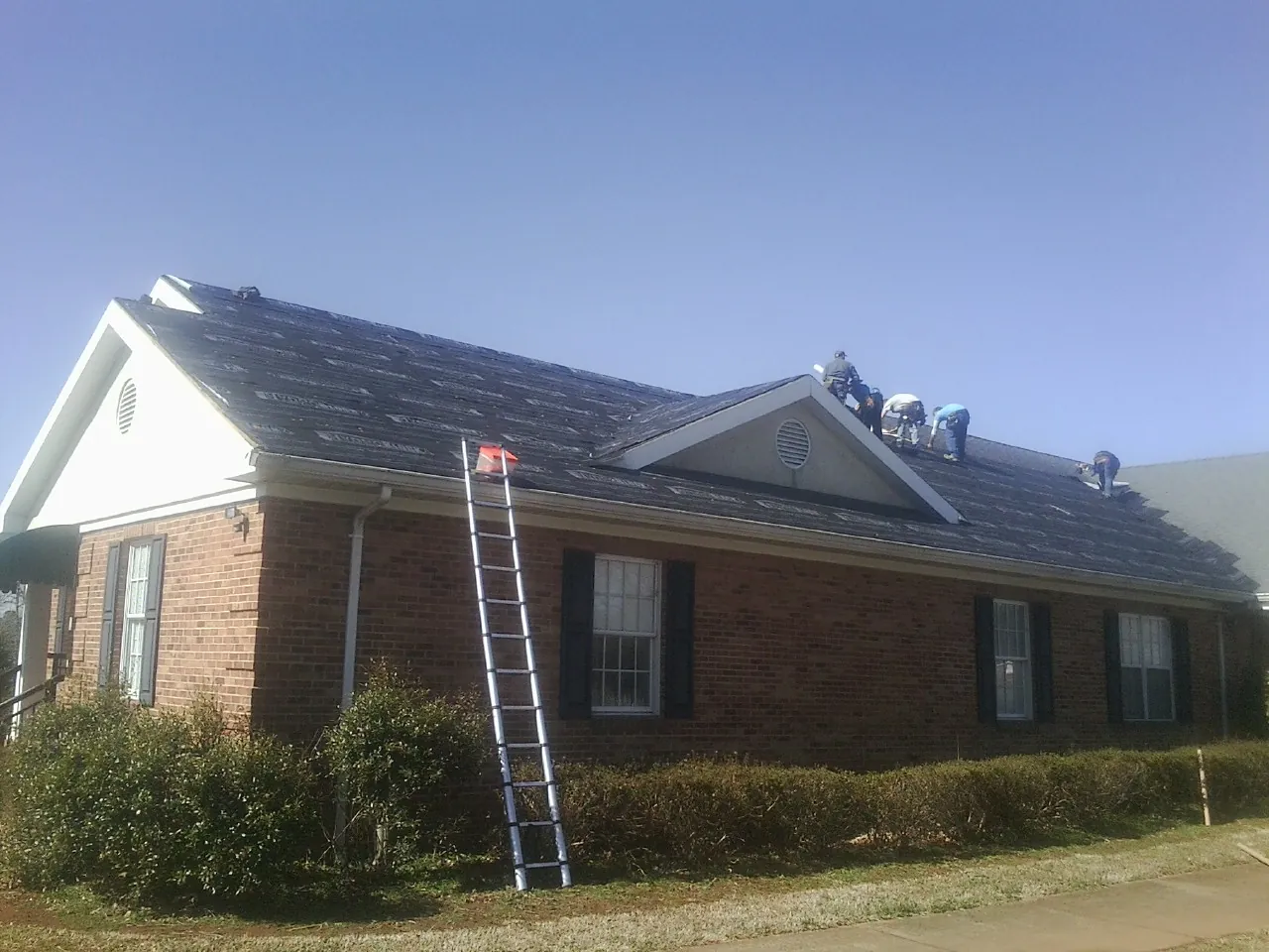 Crew tearing shingles off a brick ranch roof with a ladder set — Emergency Roof Repair work by our crew in Keene Valley