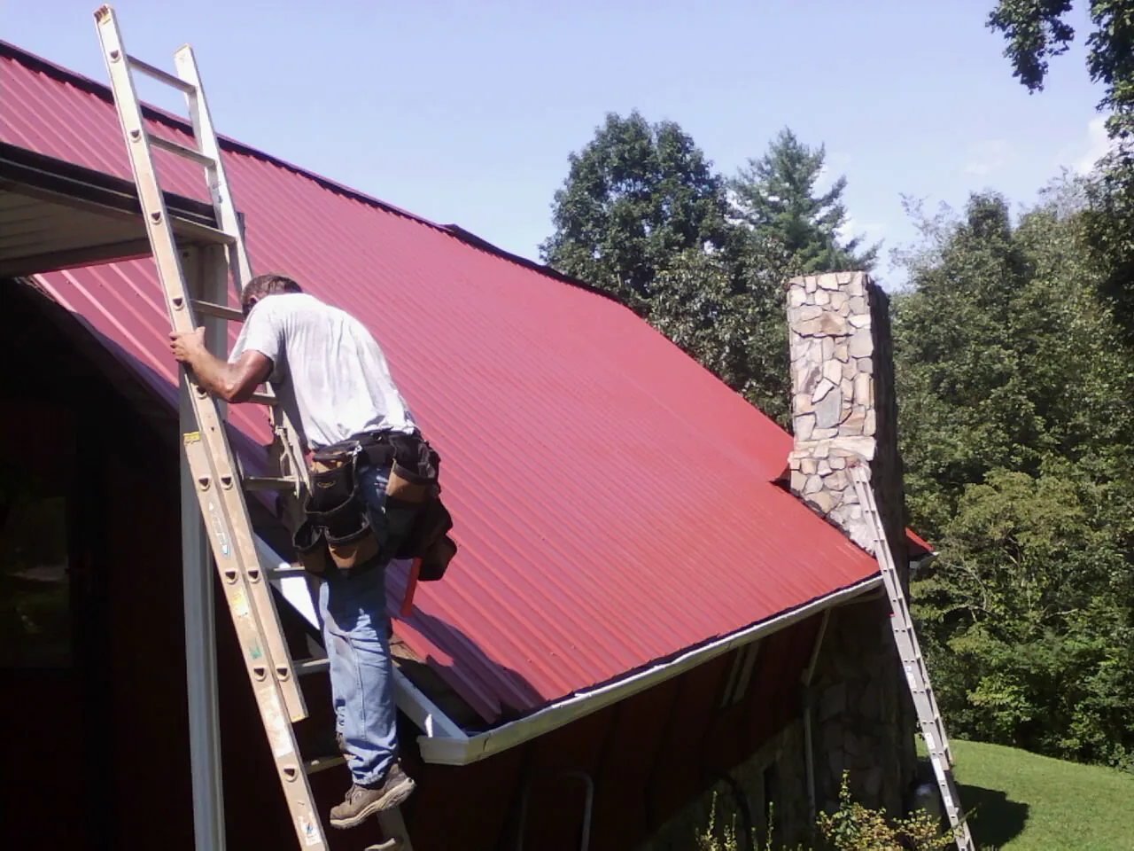 Roofer on a ladder beside a red metal roof and stone chimney — Soffit & Fascia Repair by Roofing Brothers in Keene Valley